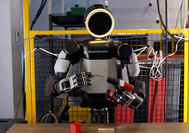 Humanoidní robot Atlas drží vrtačku a kovový nástroj, na hrudi nápis Boston Dynamics.