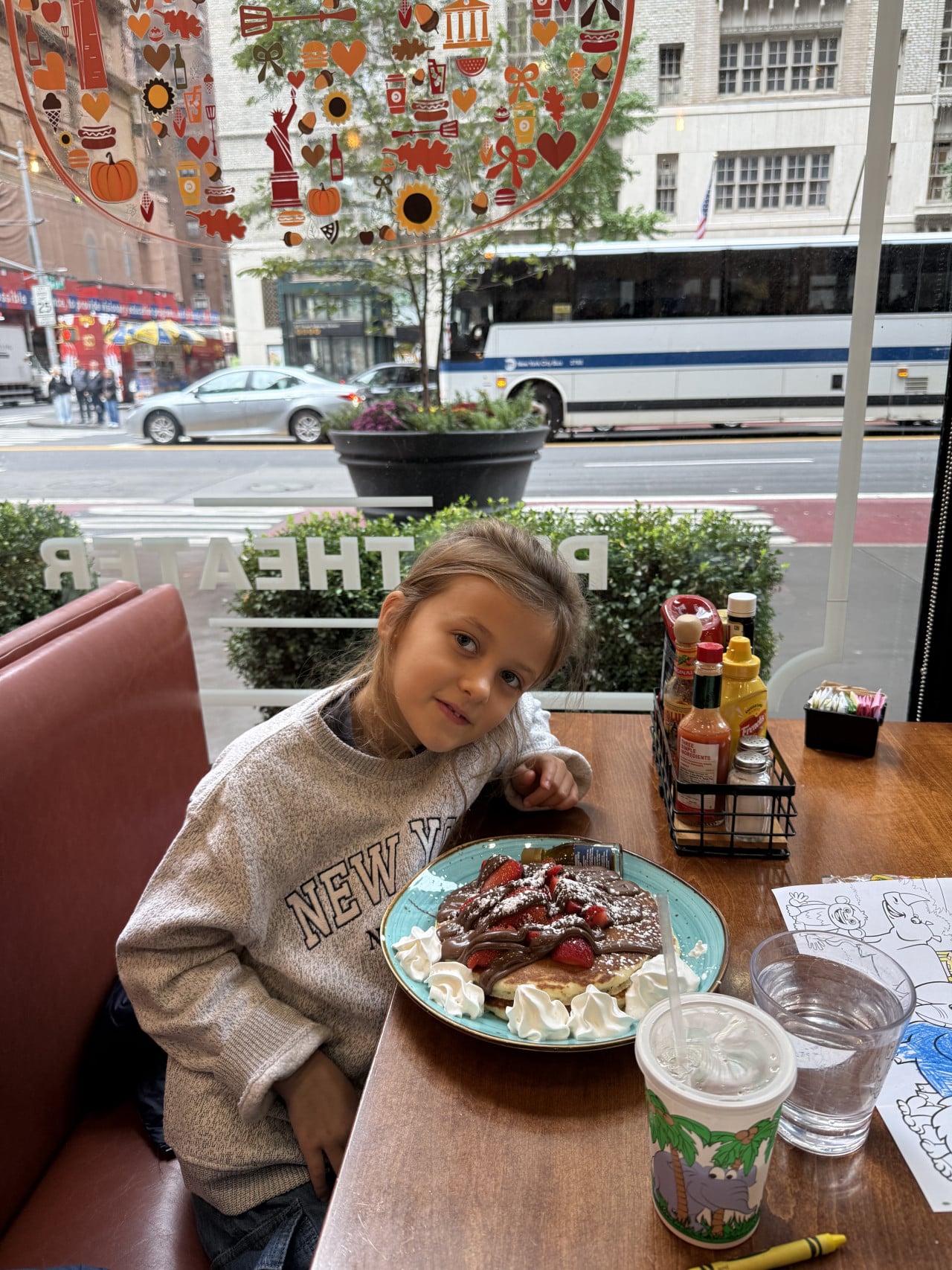 Dívka u stolu s palačinkami v newyorské restauraci, za oknem projíždí autobus.