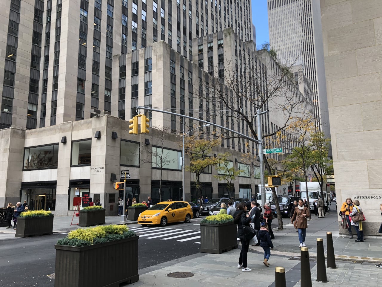 Newyorská ulice u Rockefeller Plaza s taxíkem, chodci a vysokými budovami.