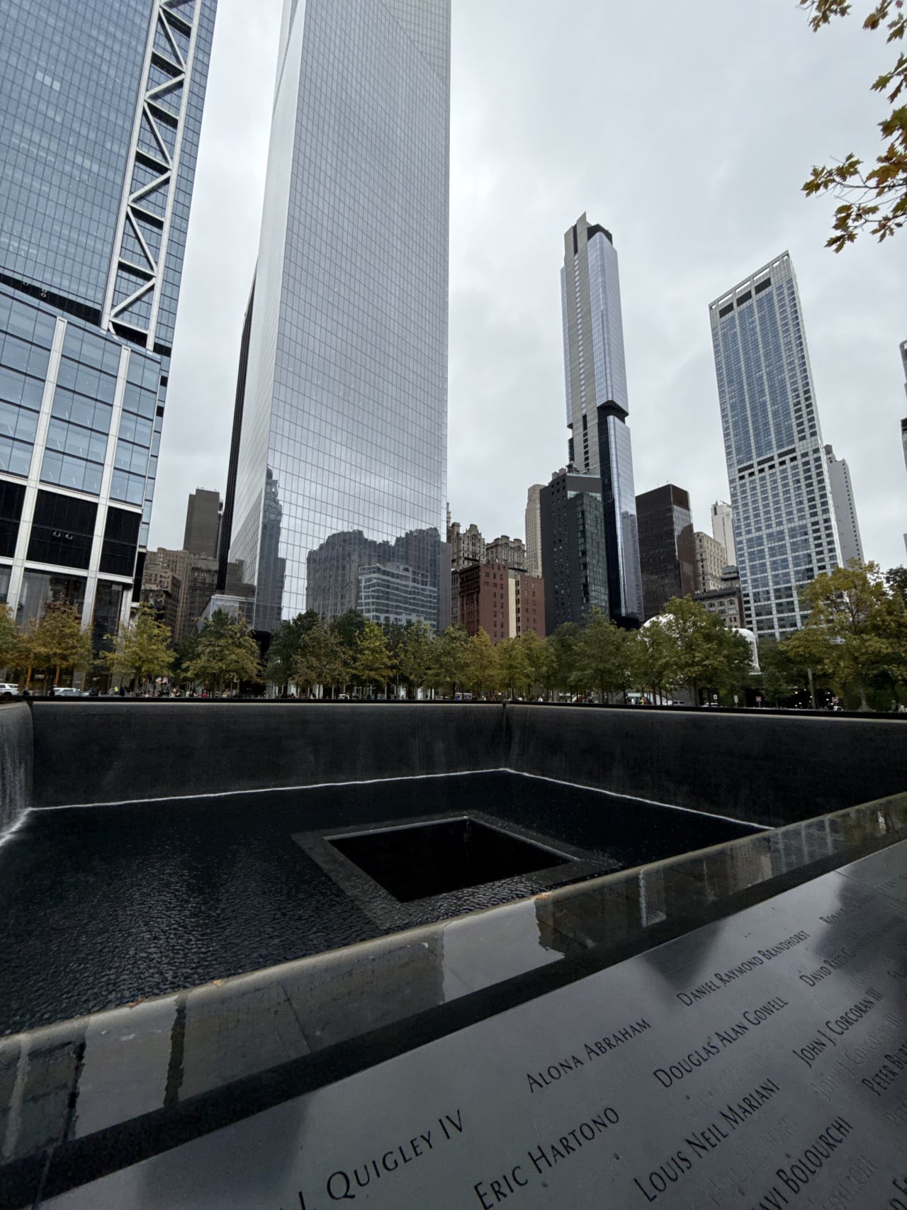 9/11 Memorial s věžemi One World Trade Center v pozadí a vyrytými jmény obětí.