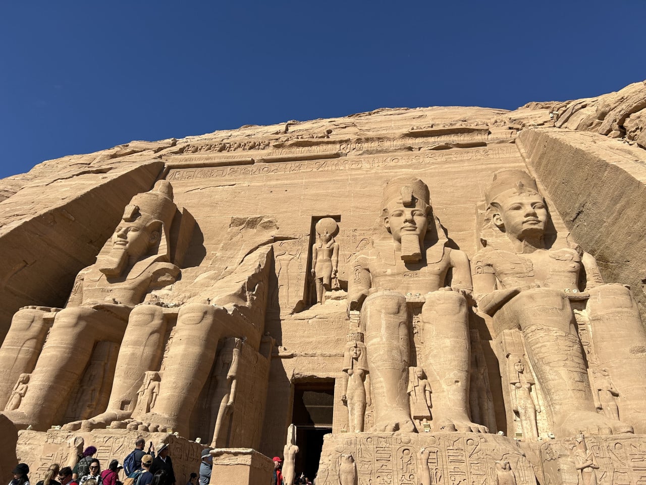 Fasáda chrámu Abu Simbel se čtyřmi sochami faraona a turisty dole, modrá obloha.