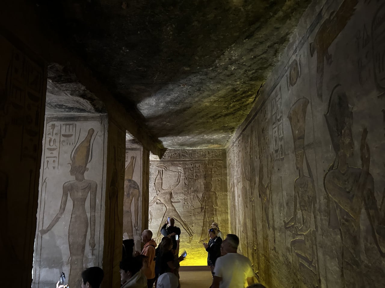Temná chrámová chodba s egyptskými reliéfy a turisty u stěn