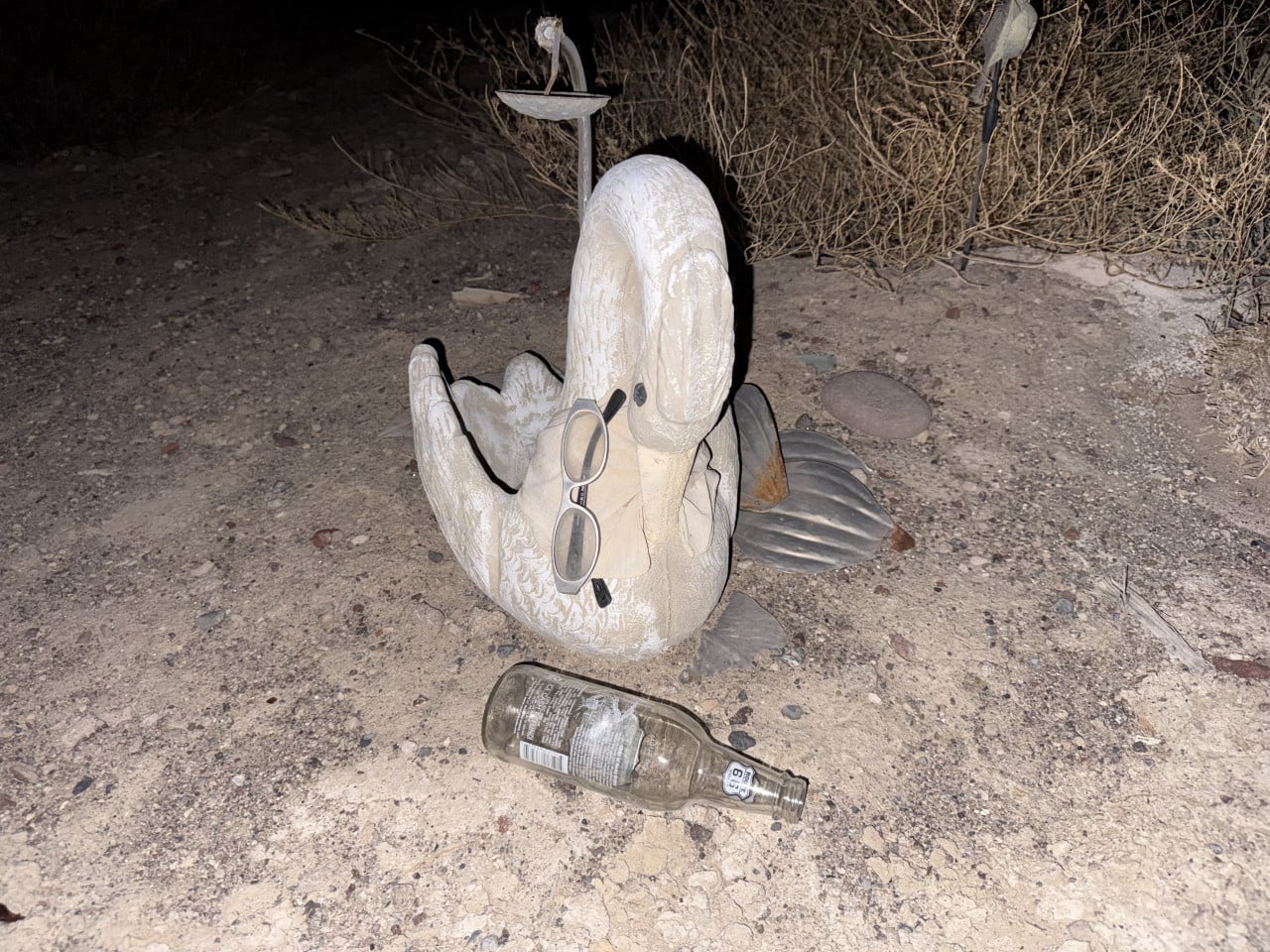 Bílá socha ležící postavy s brýlemi a lahví na kamenité zemi v noci.