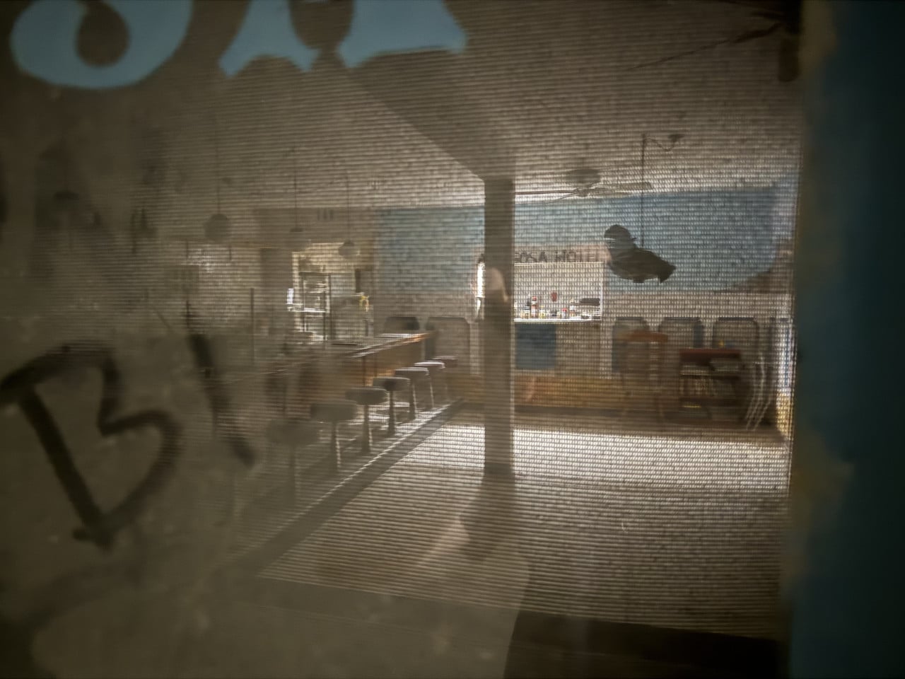 Pohled skrz síťovinu do opuštěného baru s pultem a stoličkami.