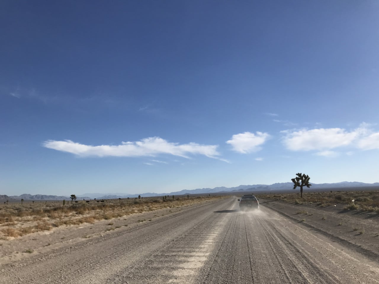 Prázdná pouštní silnice v Nevadě, auto zvedá prach pod modrou oblohou.