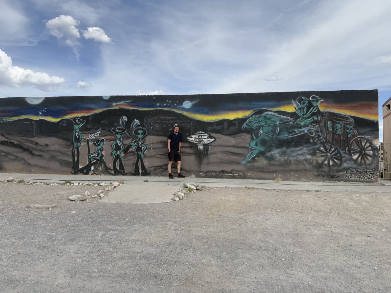 Muž stojí před muralem s mimozemšťany, UFO a kočárem v nevadské poušti.