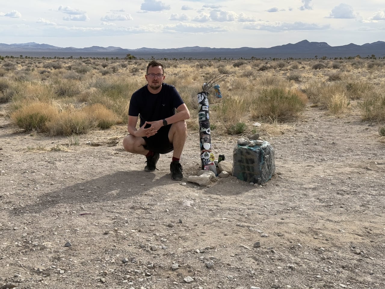 Muž v poušti u sloupku polepeného samolepkami, hory v pozadí.