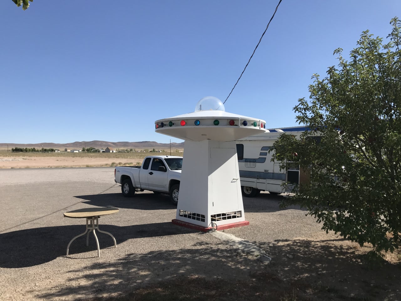 Bílá maketa UFO, pickup a obytné auto u silnice v pouštní krajině.