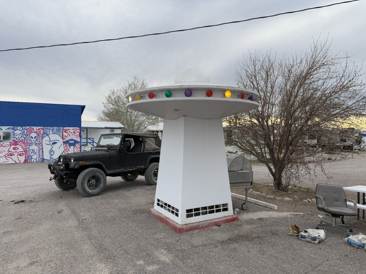 Bílý model UFO na dvoře, vedle černého Jeepu a malby mimozemšťanů na zdi.