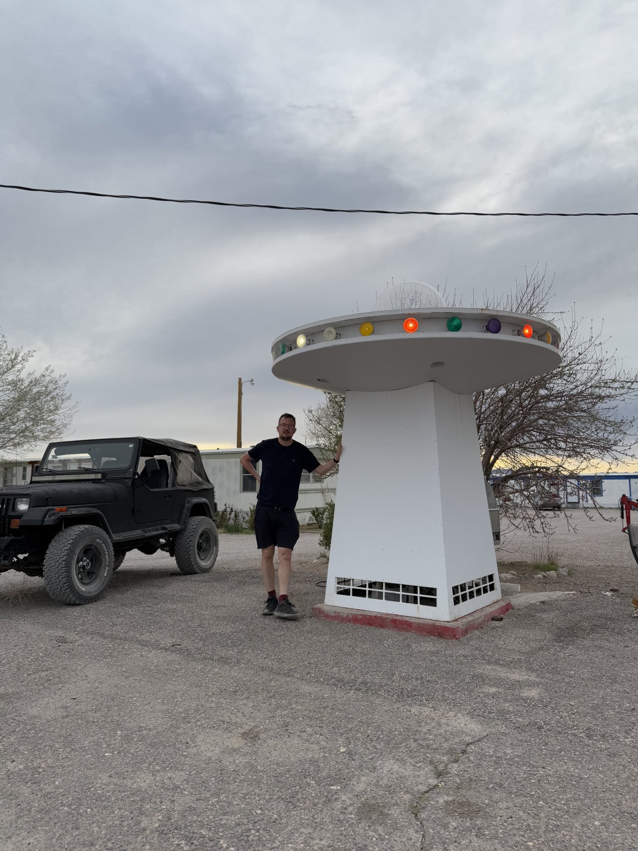 Muž stojí u modelu UFO, vedle černého terénního Jeepu, pod zataženou oblohou.
