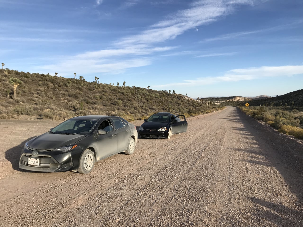 Dvě auta na prašné cestě v nevadské poušti, kolem nízké kopce a modrá obloha.