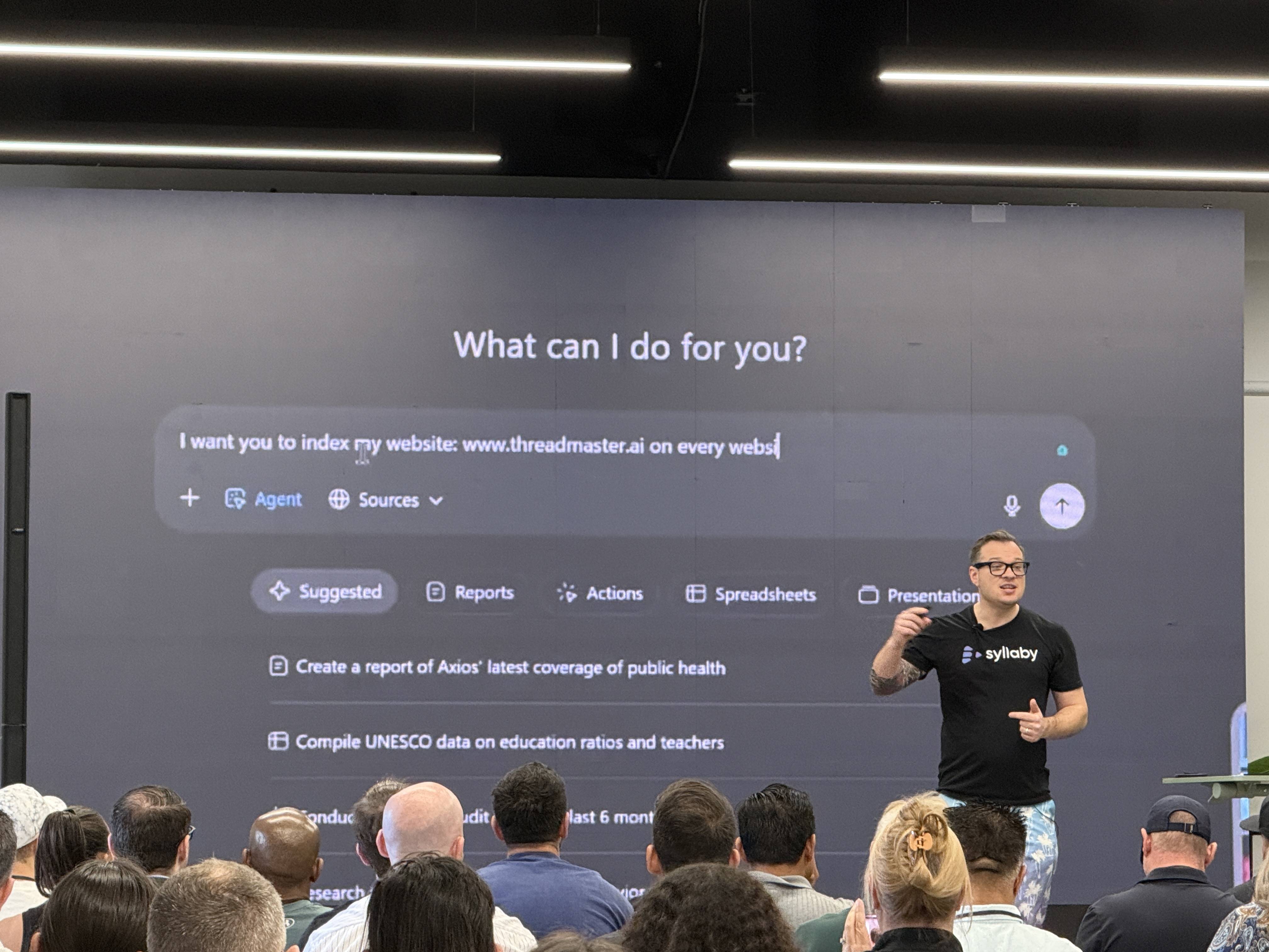 Přednášející v tričku Syllaby před publikem, na plátně What can I do for you? www.threadmaster.ai