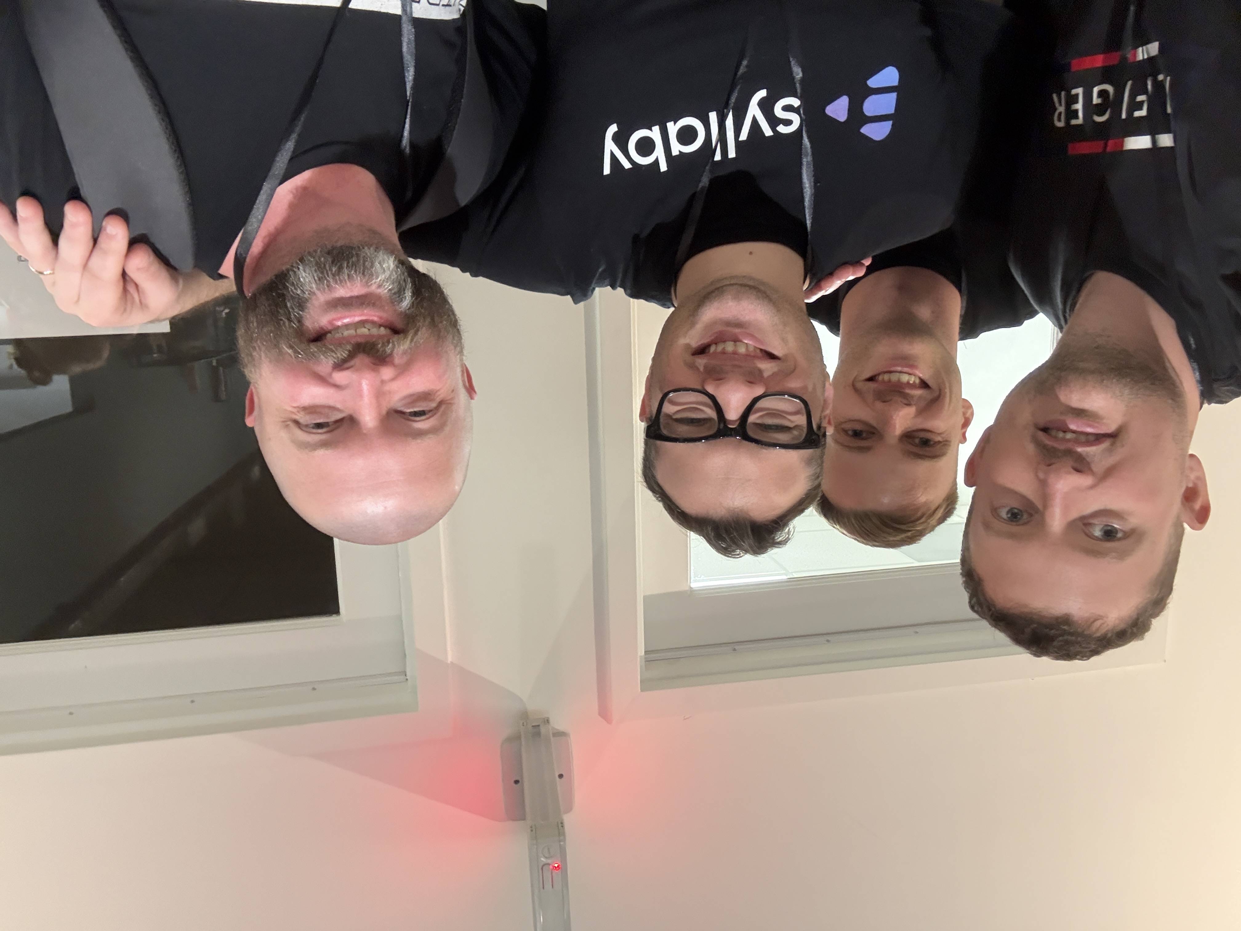 Selfie čtyř usmívajících se mužů v interiéru; trička s nápisy syllaby a HILFIGER.