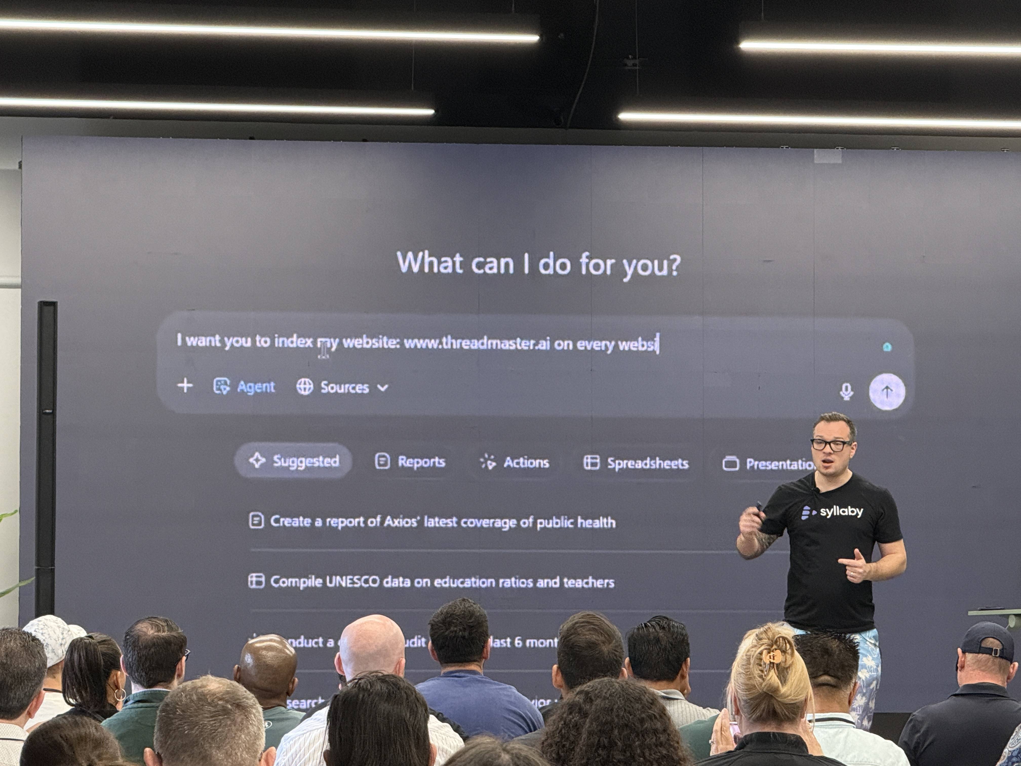 Prezentující muž v tričku Syllaby před plátnem s textem What can I do for you?