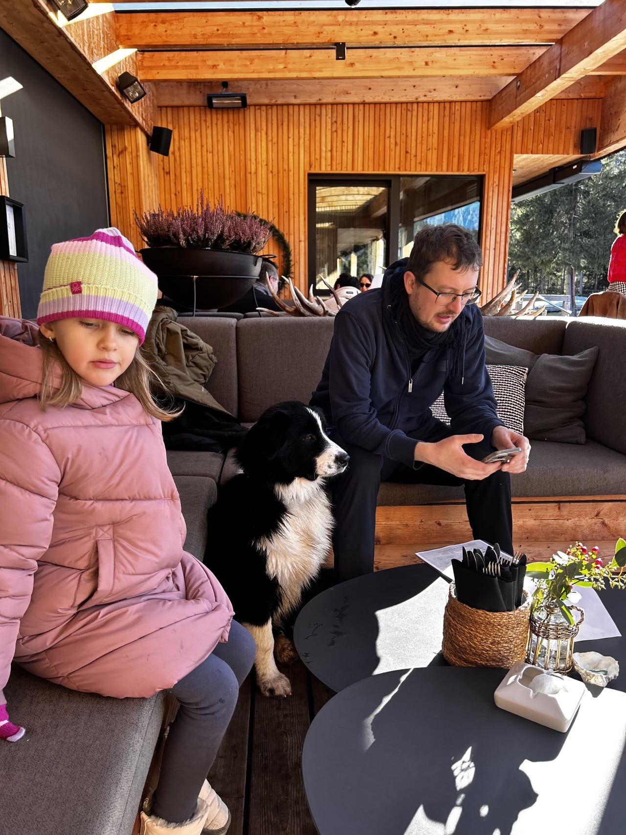 Dívka v růžové bundě, pes a muž s mobilem na dřevěné terase.