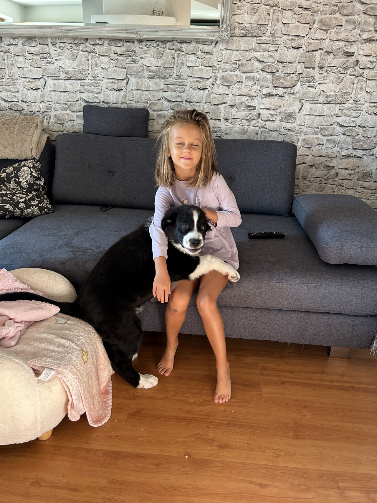 Dívka sedí na šedé sedačce a objímá černobílého psa v obýváku.