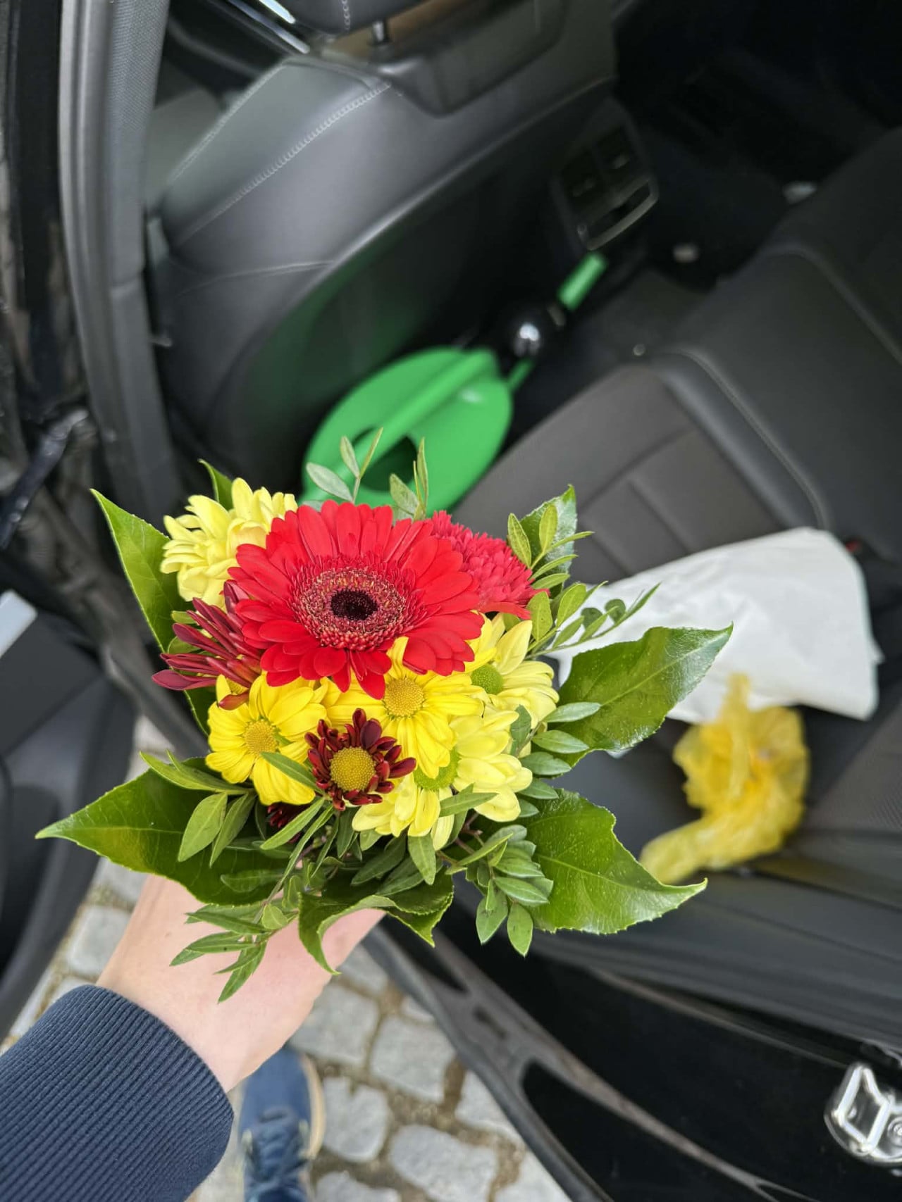 Ruka drží barevnou kytici gerber a chryzantém u otevřených dveří auta.