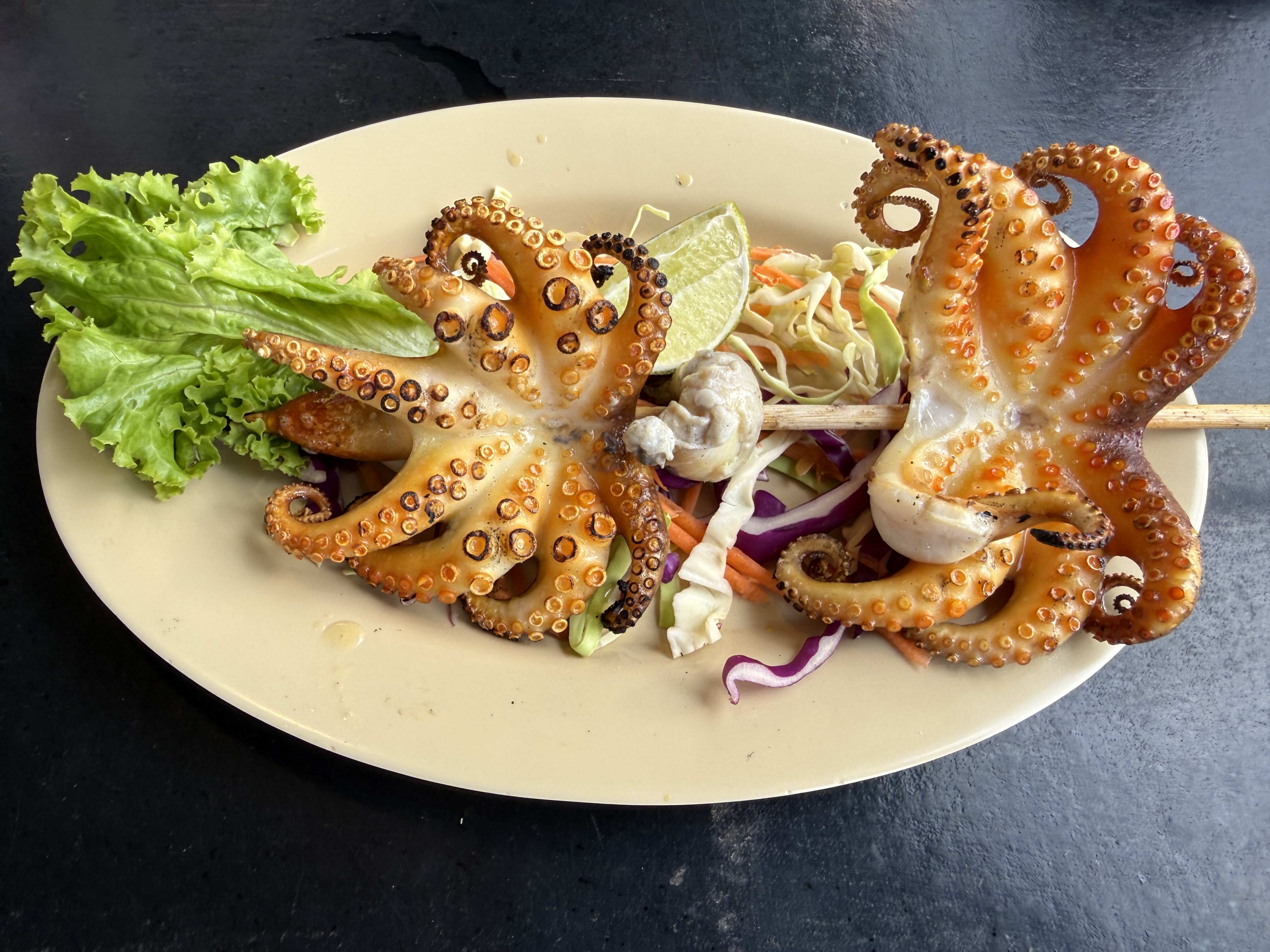 Dvě grilované chobotnice na špejli, salátový mix, limetka a list salátu na talíři