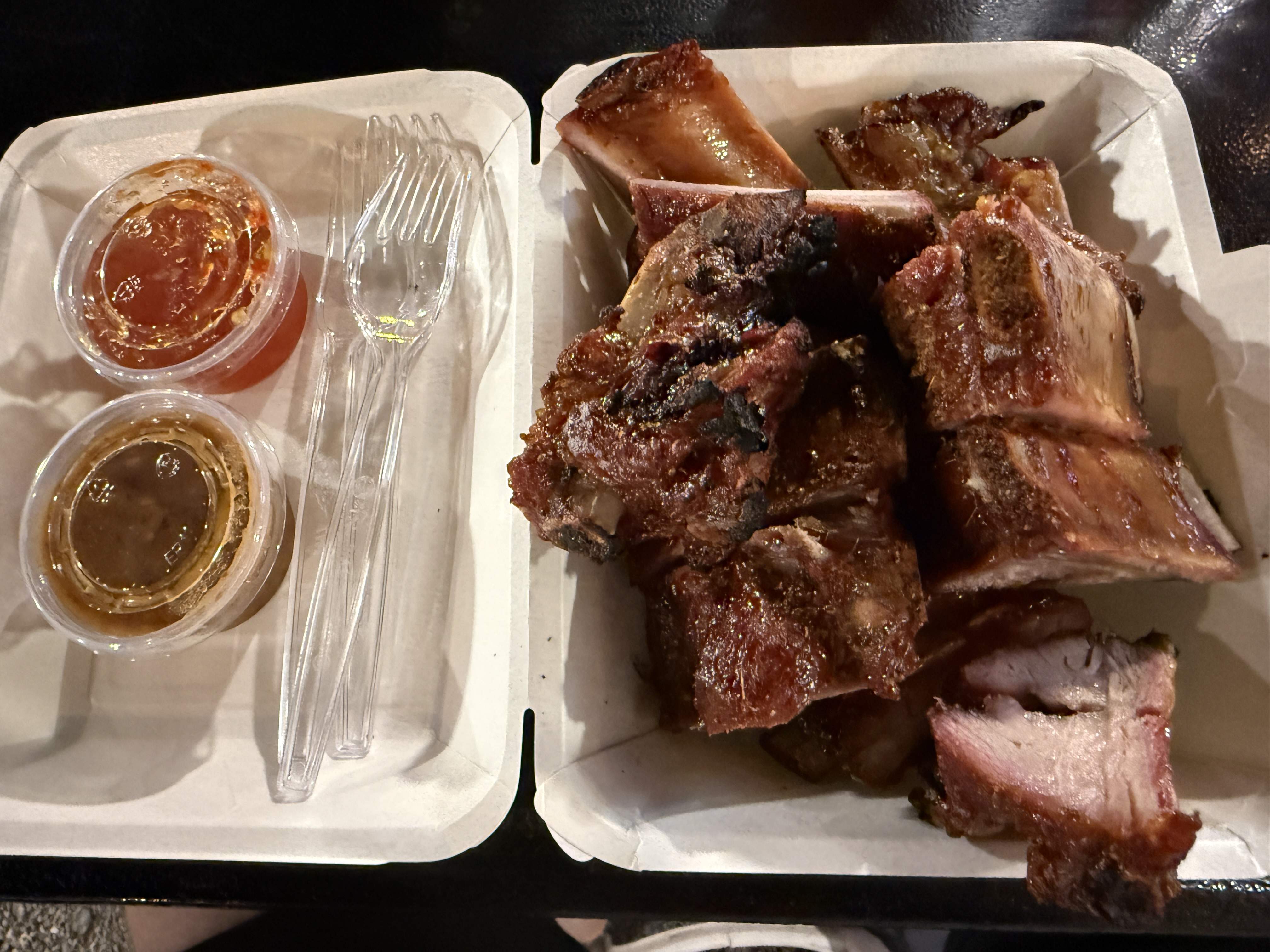 Porce grilovaných vepřových žeber v krabičce, vedle omáčky a plastové příbory