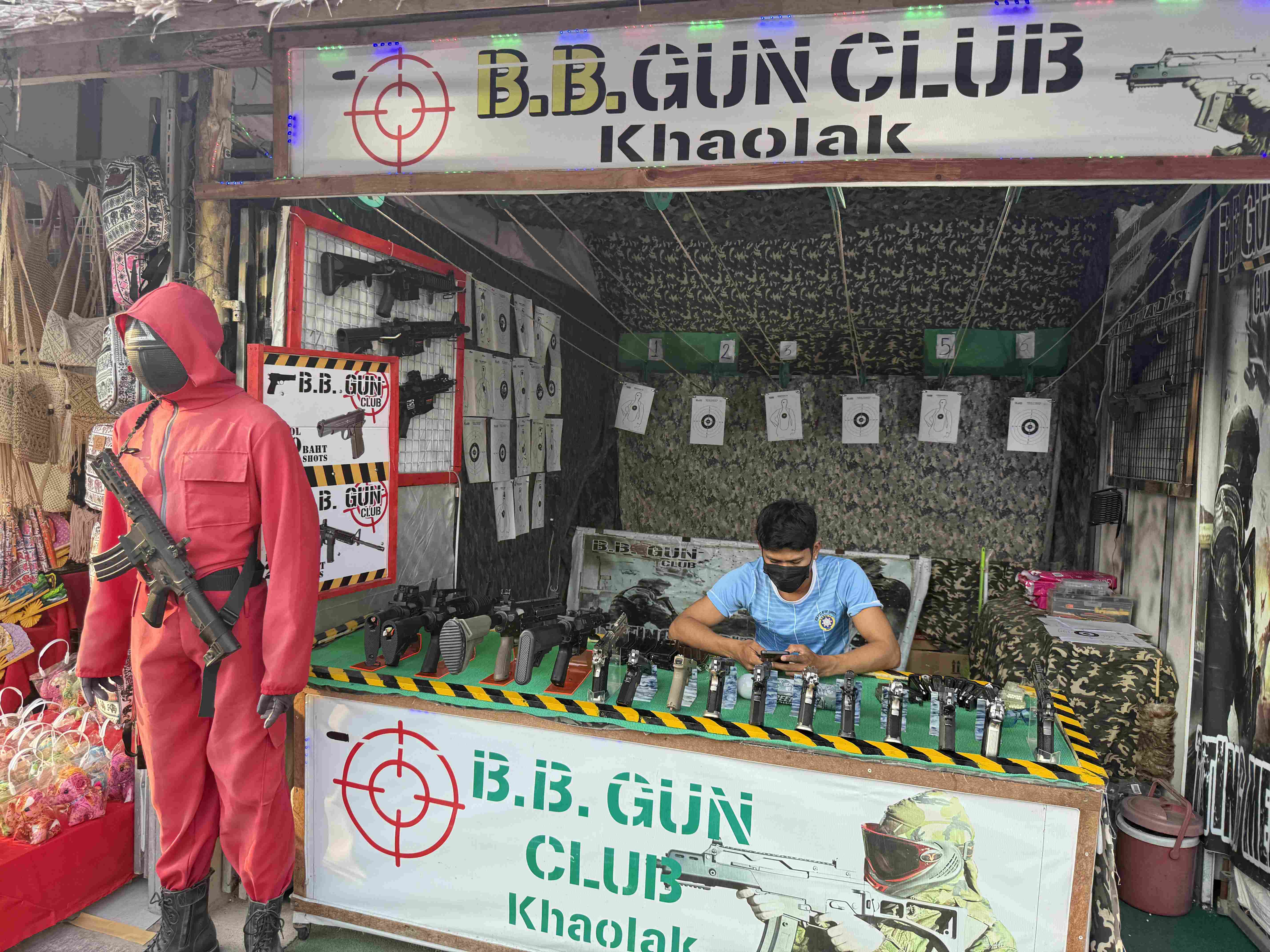 Stánek B.B. GUN CLUB Khaolak, figurína v červeném obleku a zbraně na stole