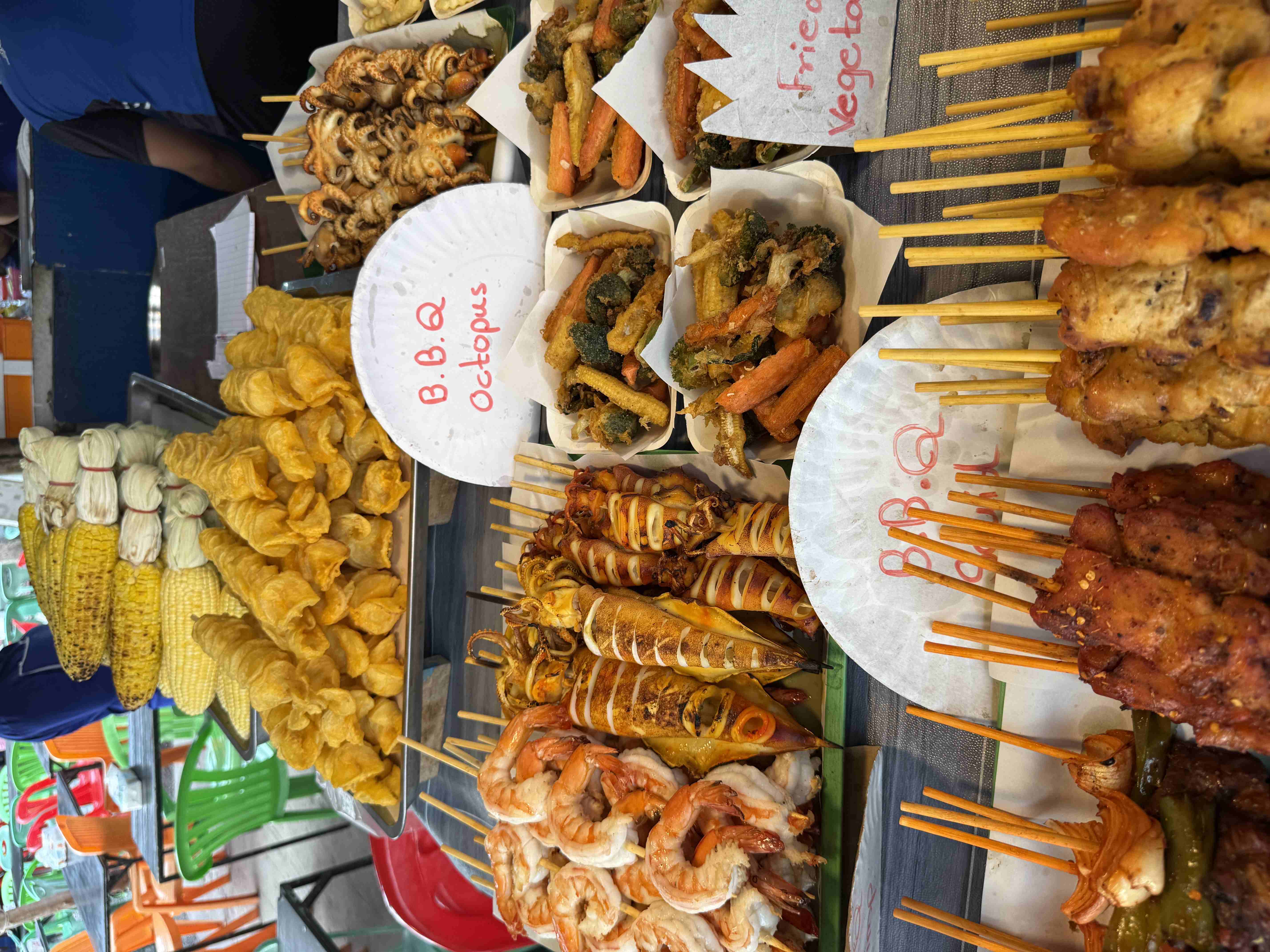 Stánek s grilovanými špízy, kukuřicí a cedulí B.B.Q Octopus, Fried Vegetables.