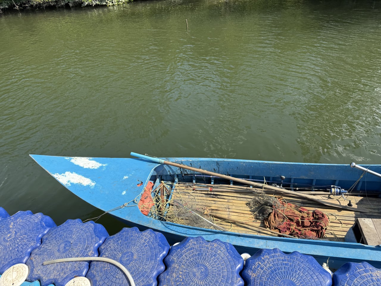 Modrý dřevěný člun s pádlem a rybářskými sítěmi u modrého plastového mola