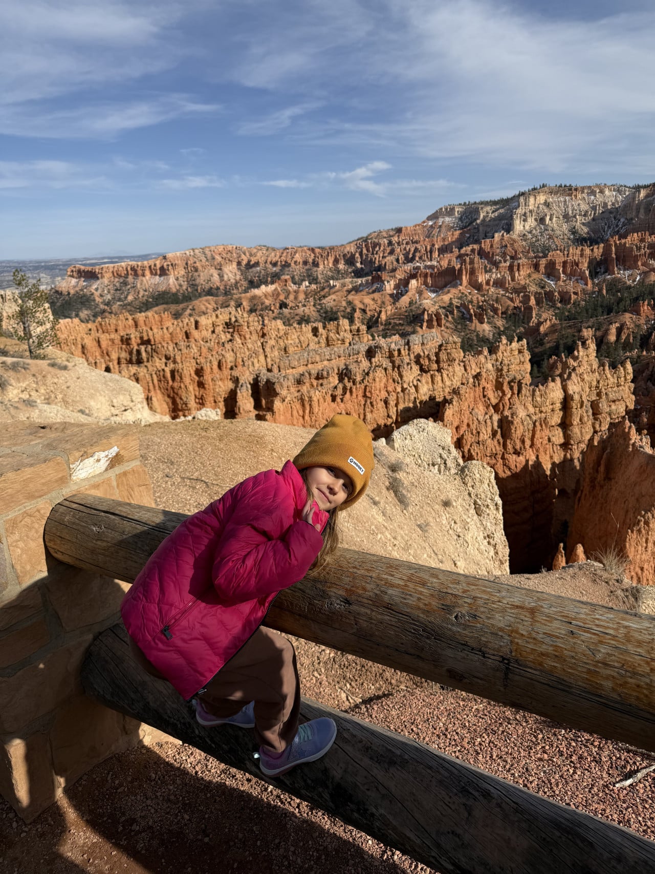 Dítě v růžové bundě u dřevěného zábradlí, za ním Bryce Canyon hoodoos