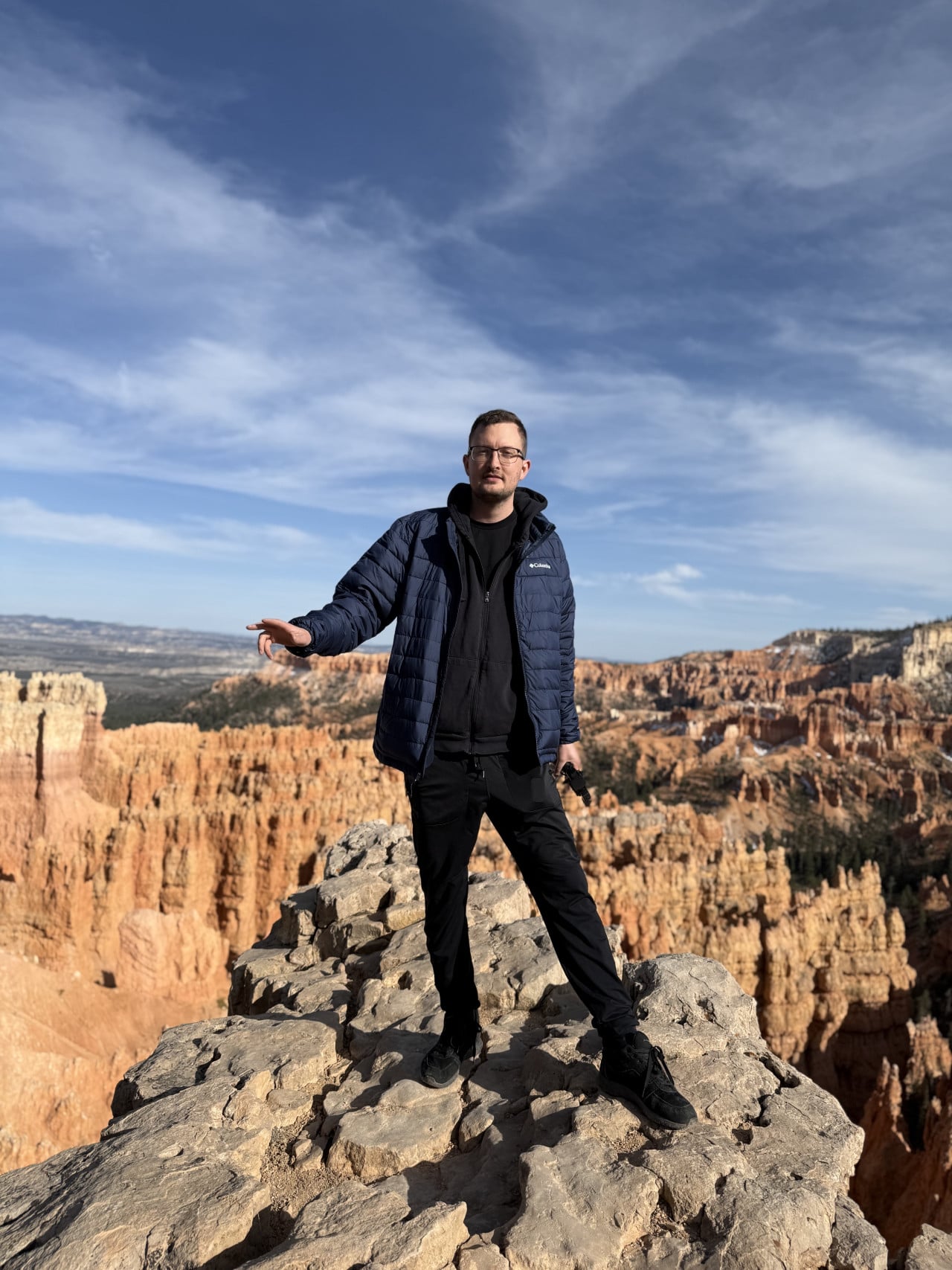 Muž stojí na skále, za ním oranžové věže Bryce Canyonu a modrá obloha.