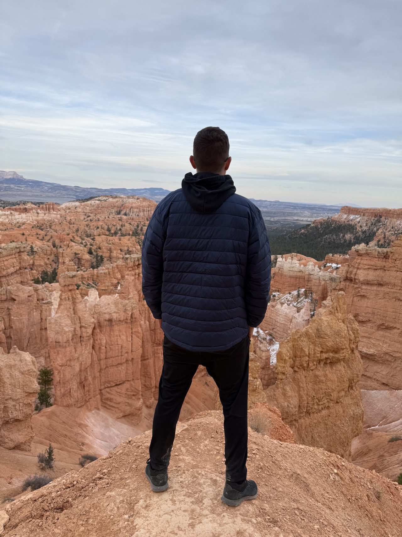 Muž v bundě stojí na okraji útesu, výhled na hoodoo věže Bryce Canyonu