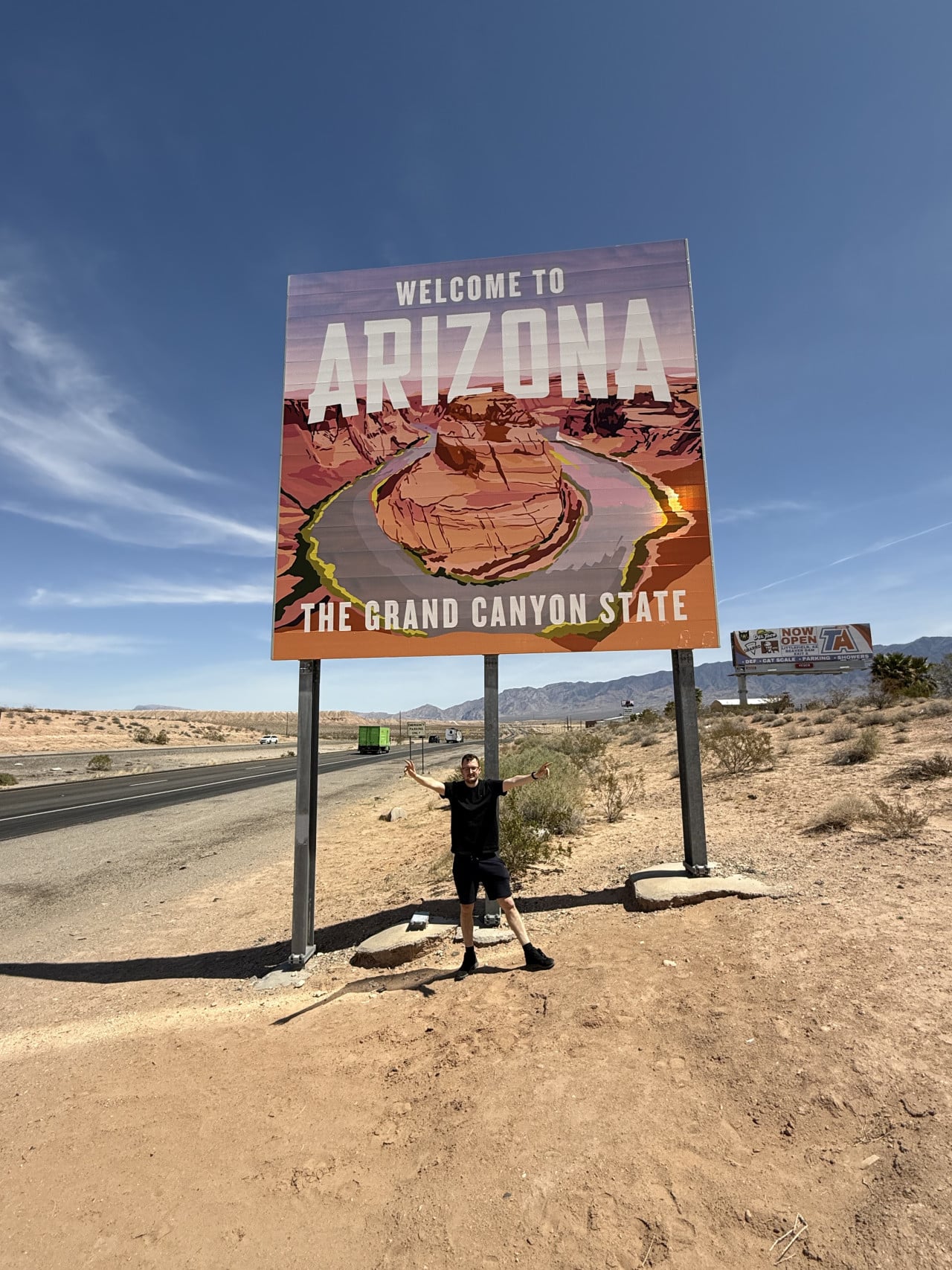 Muž pózuje pod cedulí WELCOME TO ARIZONA, THE GRAND CANYON STATE u silnice.