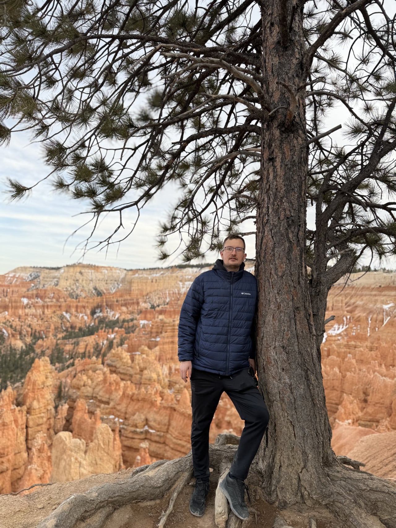 Muž v modré bundě opřený o borovici, v pozadí hoodoo Bryce Canyonu.