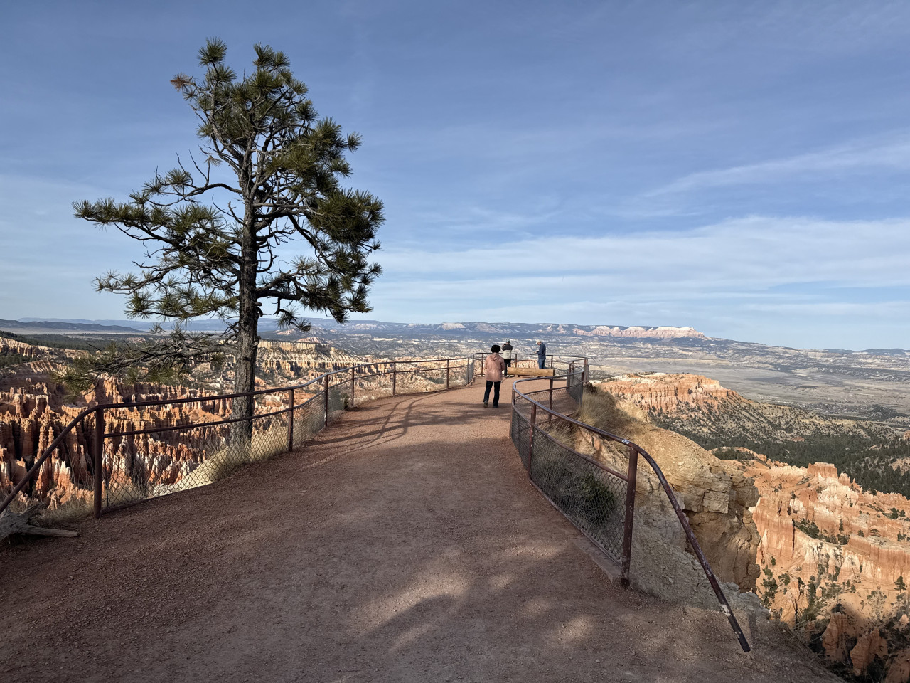 Vyhlídková stezka s zábradlím, borovice a lidé nad skalními věžemi Bryce Canyonu.