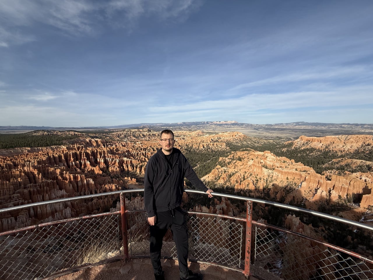 Turista na vyhlídce s kovovým zábradlím, za ním oranžové věže Bryce Canyonu