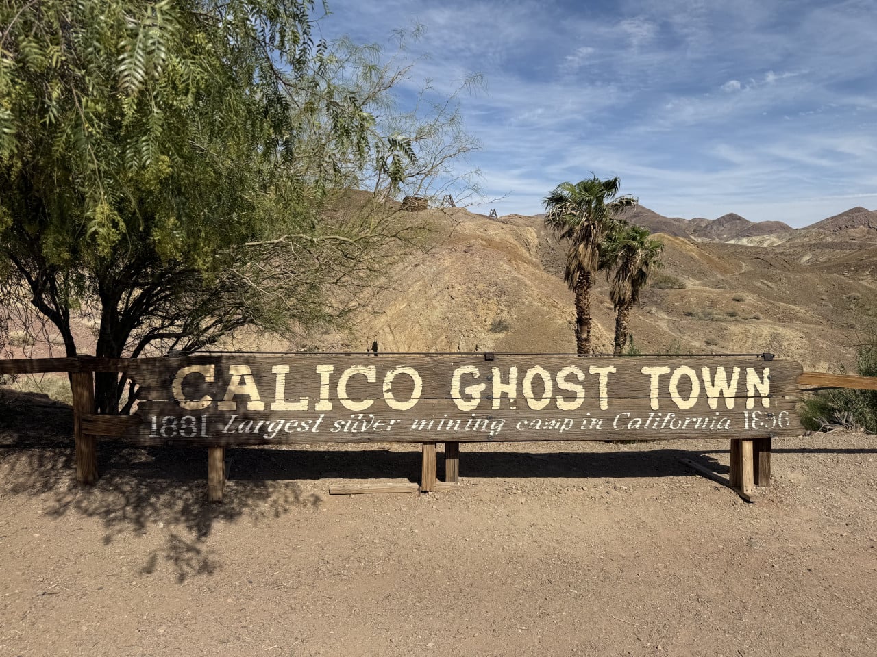 Dřevěná cedule Calico Ghost Town v pouštní krajině s palmami a kopci