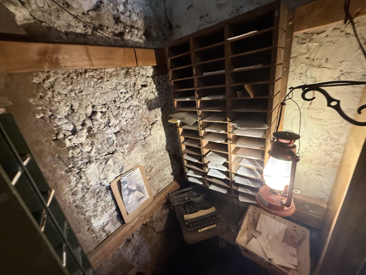 Interiér staré poštovní místnosti s přihrádkami, psacím strojem a petrolejovou lampou v kamenné stěně