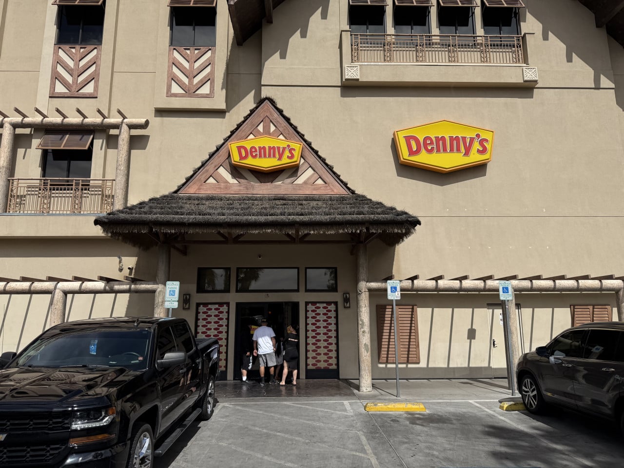 Před restaurací Denny’s stojí zaparkovaná auta a lidé u vchodu.