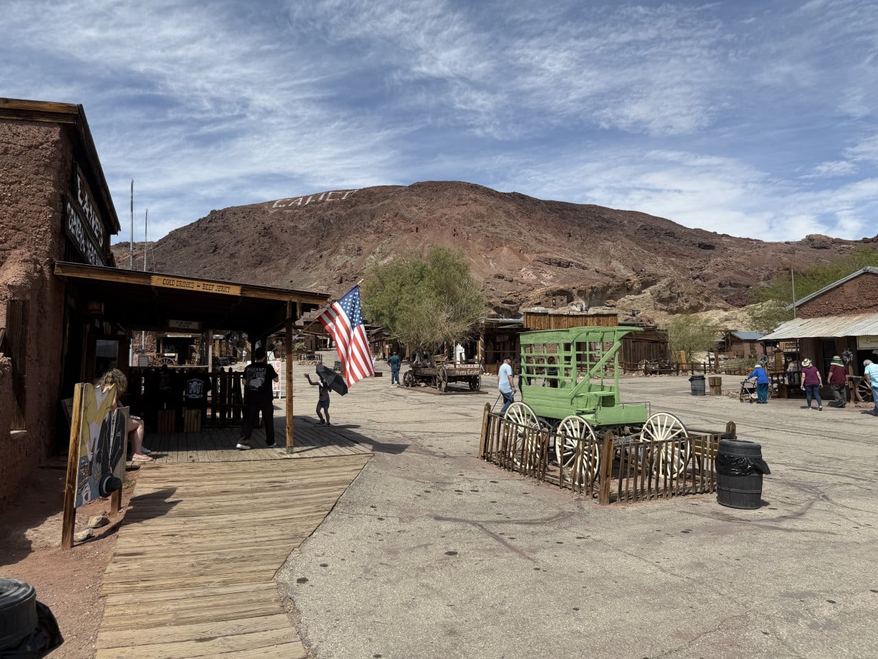 Pohled na Calico Ghost Town s americkou vlajkou, dřevěnými budovami a pouštním kopcem v pozadí