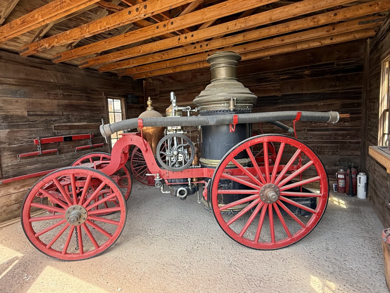 Stará hasičská parní stříkačka s červenými koly v dřevěné budově.