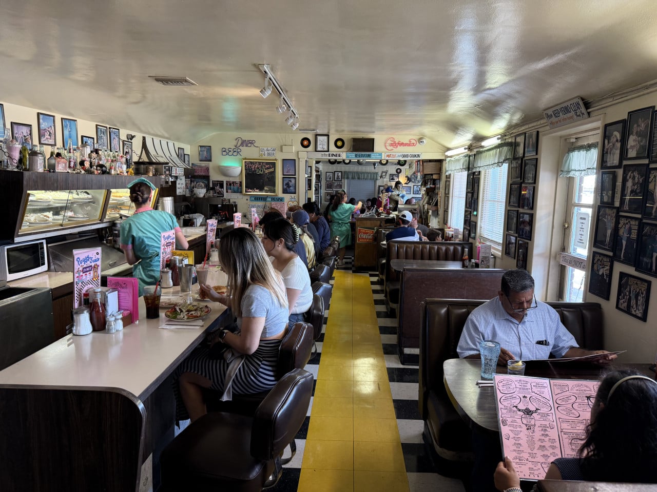 Rušný americký diner s hosty, servírkami, vitrinou s dorty a žlutou podlahovou uličkou