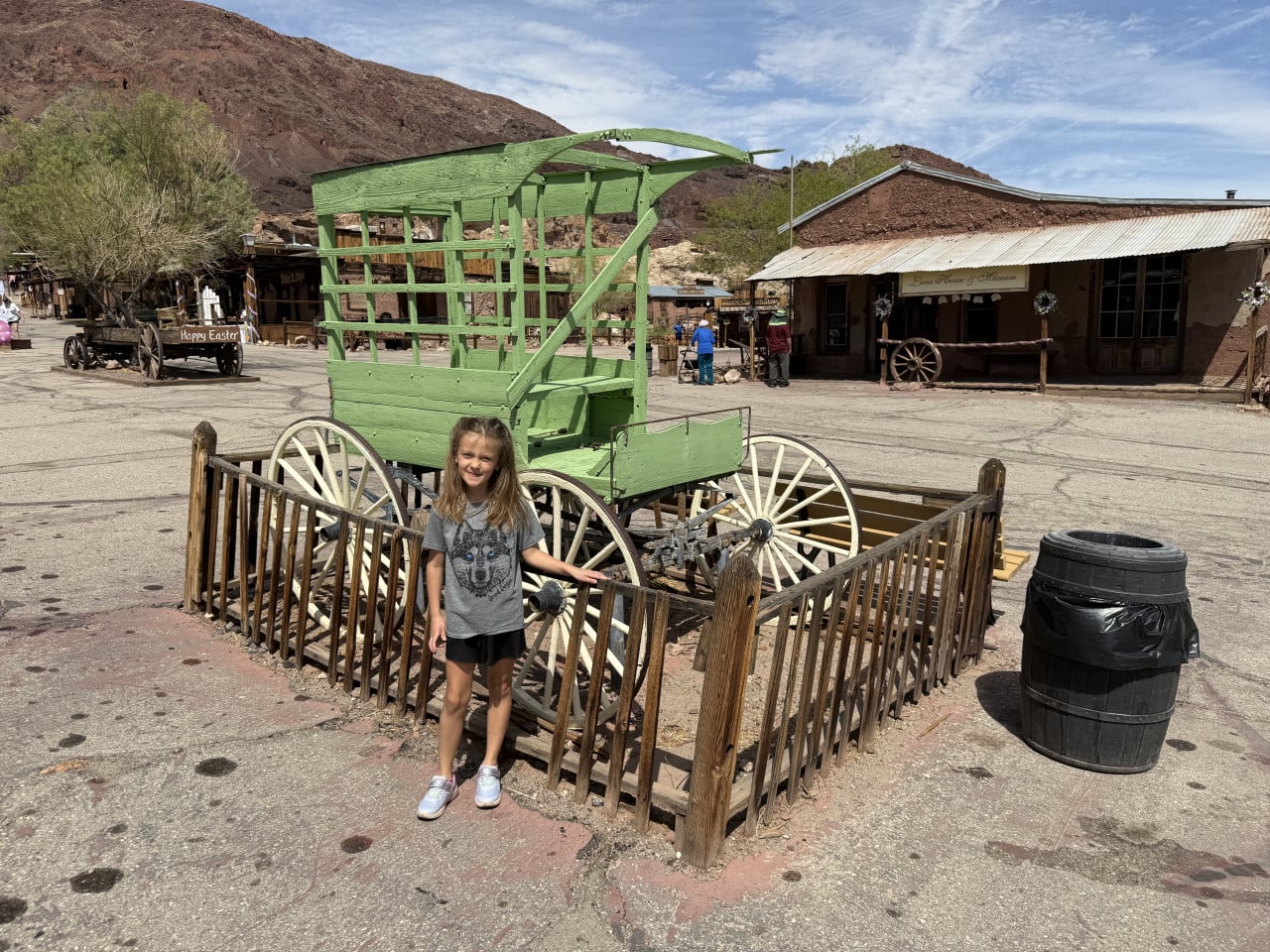 Dívka stojí u zeleného dřevěného vozu v Calico Ghost Town mezi pouštními budovami.