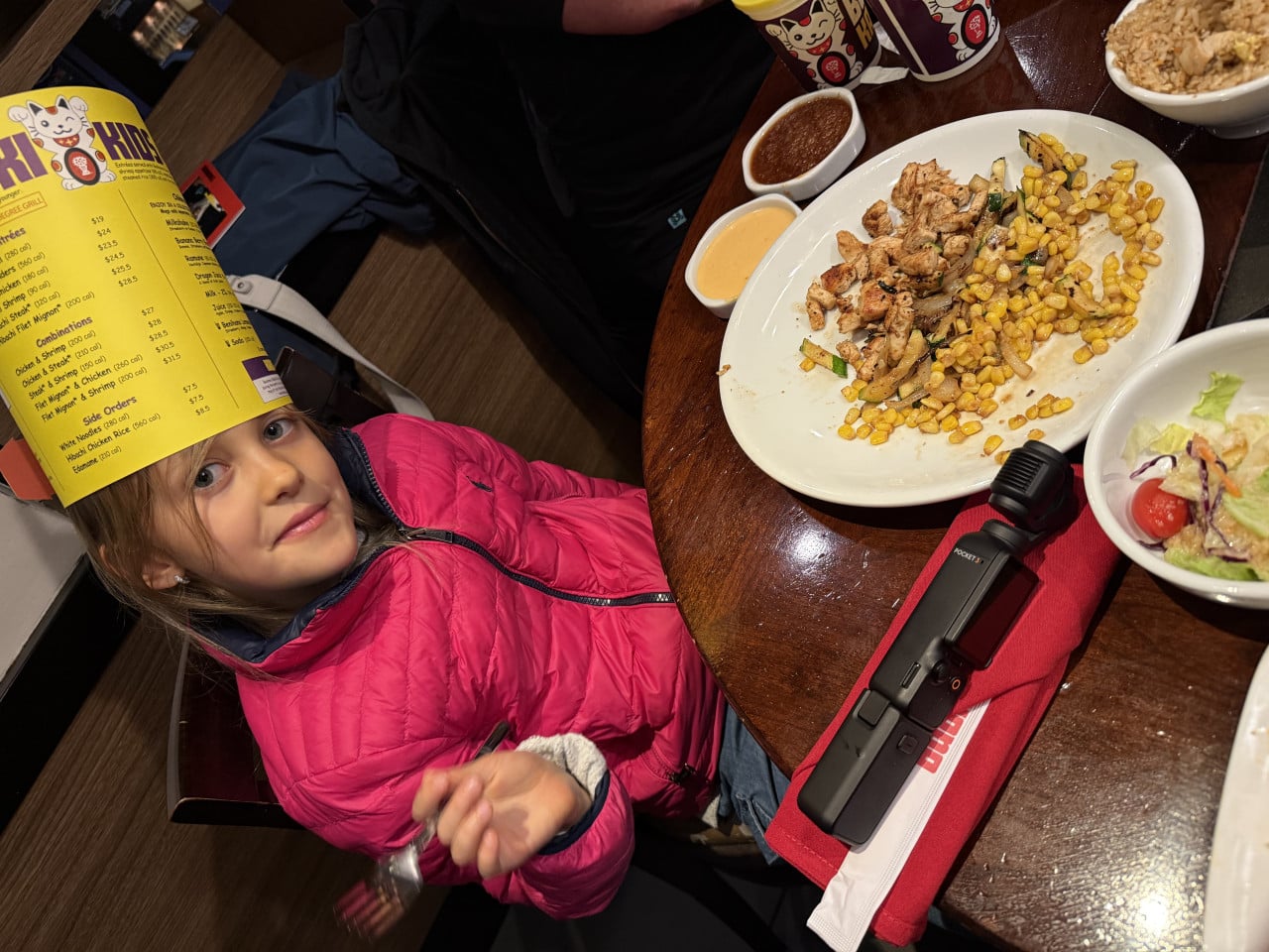 Dívka v růžové bundě jí v restauraci Benihana, před sebou má talíř s kuřetem a kukuřicí.