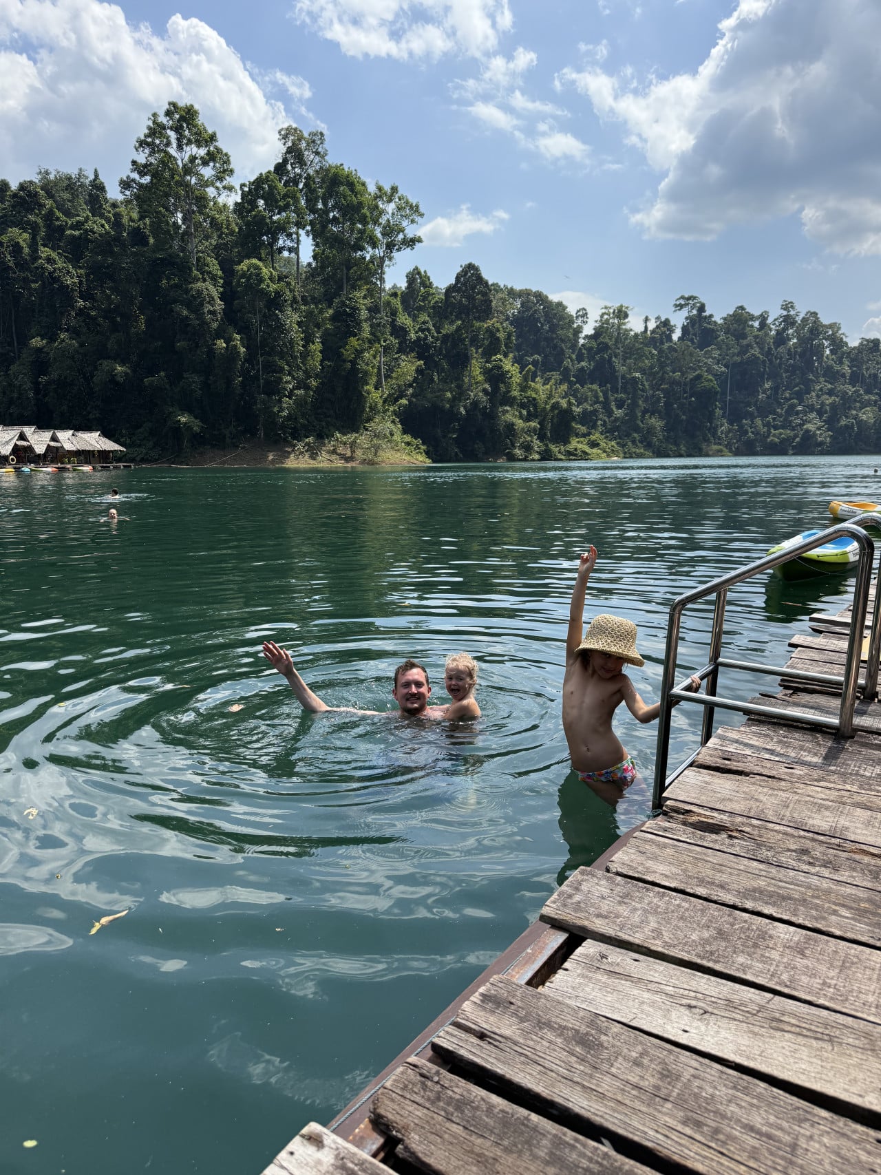 Dítě v klobouku mává z mola, rodina plave v jezeře, džungle v pozadí