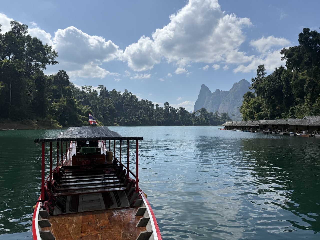 Dřevěná loď s thajskou vlajkou na jezeře, vpravo plovoucí bungalovy, džungle a skály.