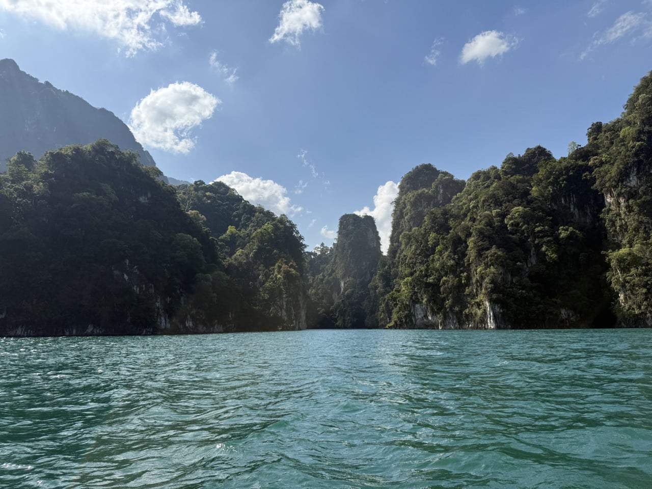 Smaragdová hladina jezera Cheow Lan obklopená zalesněnými vápencovými skalami a modrou oblohou
