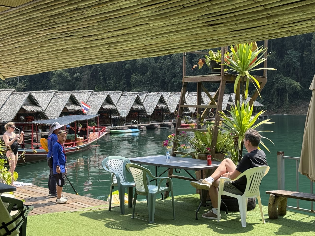 Muži a turisté na plovoucím molu, longtail loď a bungalovy na jezeře