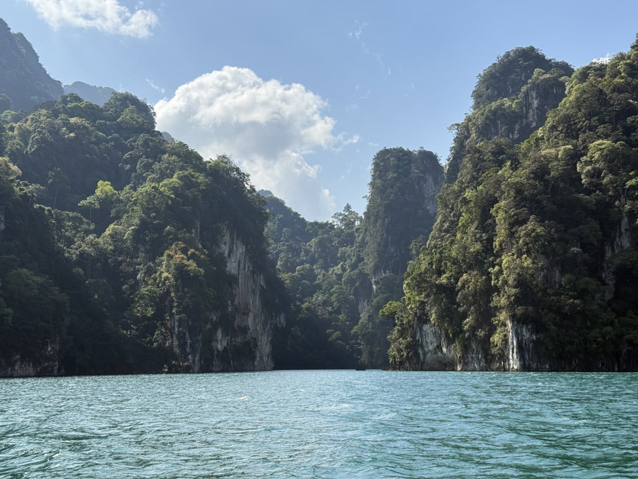 Tyrkysová hladina Cheow Lan Lake obklopená zalesněnými vápencovými skalami, modrá obloha.