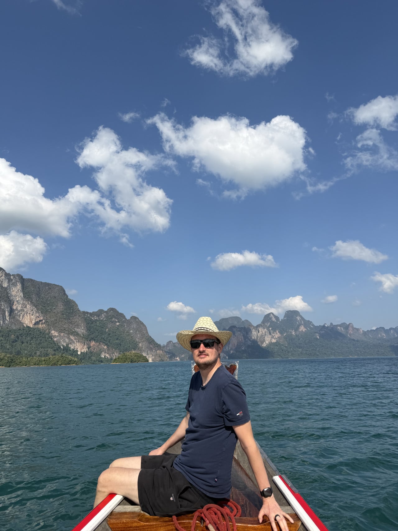 Muž v klobouku sedí na přídi lodi, v pozadí vápencové hory a jezero