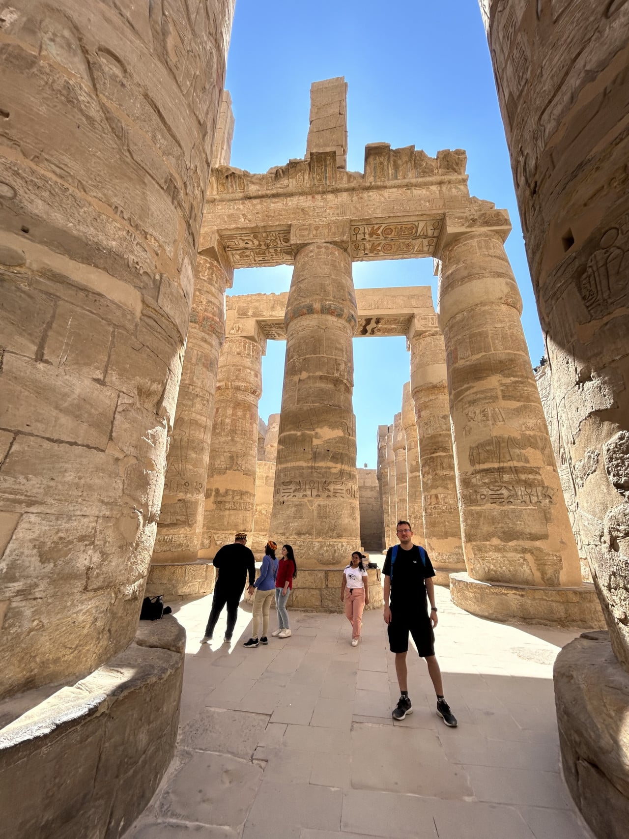 Turisté mezi obřími sloupy chrámu Karnak pod modrou oblohou