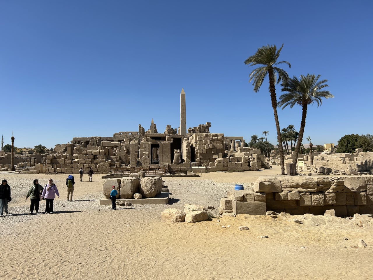 Ruiny chrámu Karnak s obeliskem, palmami a turisty na písčitém nádvoří