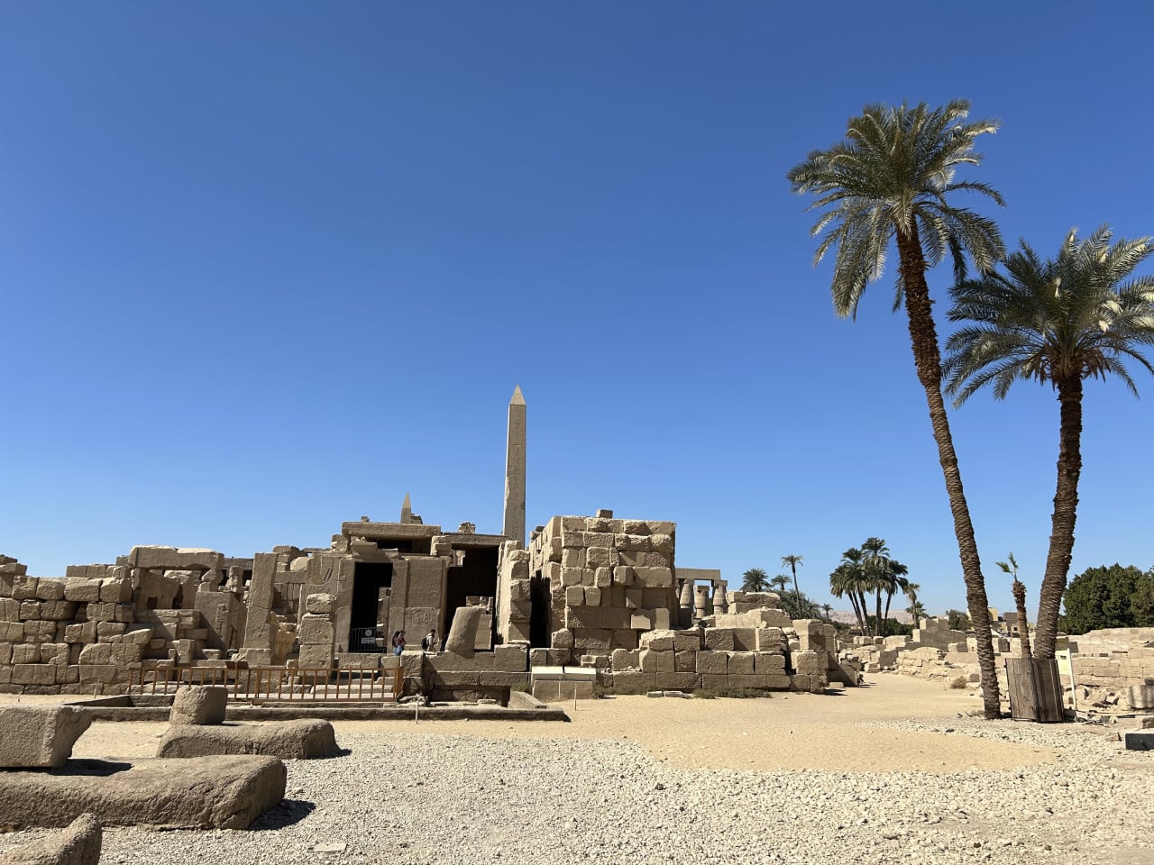 Ruiny chrámu Karnak s obeliskem a palmami pod modrou oblohou